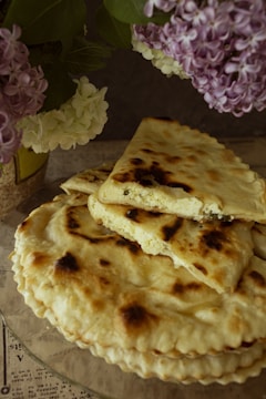 A stack of golden-brown flatbreads sits on a glass plate, with one cut into triangles, revealing a cheese filling. Surrounding the bread are clusters of lilac and white hydrangeas, adding a floral touch.