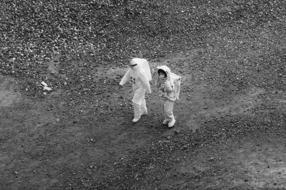 two people in space suits walking on a dirt field