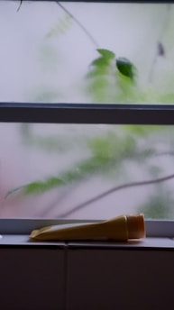A yellow tube of product lies on a tiled surface or windowsill. The background consists of a frosted glass window with a blurred view of green leaves and stems visible through it.