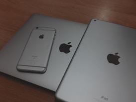 A silver iPhone is placed on top of two larger silver devices, likely a MacBook and an iPad, all featuring the Apple logo.