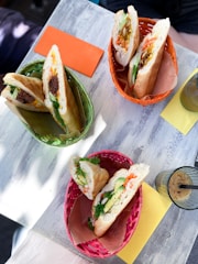 Three baskets, each containing a baguette sandwich, are placed on a light wooden table. The sandwiches are filled with various ingredients such as meat, vegetables, and herbs. Accompanying the baskets, there are colored napkins, one orange and one yellow, and a glass with a drink containing ice cubes and a straw.