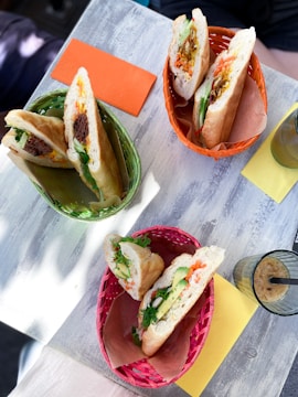 Three baskets, each containing a baguette sandwich, are placed on a light wooden table. The sandwiches are filled with various ingredients such as meat, vegetables, and herbs. Accompanying the baskets, there are colored napkins, one orange and one yellow, and a glass with a drink containing ice cubes and a straw.