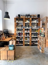 A quaint shop interior showcasing an extensive collection of bottled beverages, predominantly beer, arranged neatly on wooden shelves against a white wall. The rustic aesthetic is complemented by a large black hanging lamp, a wooden table, and crates that serve as both storage and decor. Several brown paper bags and packaging with vibrant labels are visible on the table.