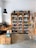 A quaint shop interior showcasing an extensive collection of bottled beverages, predominantly beer, arranged neatly on wooden shelves against a white wall. The rustic aesthetic is complemented by a large black hanging lamp, a wooden table, and crates that serve as both storage and decor. Several brown paper bags and packaging with vibrant labels are visible on the table.