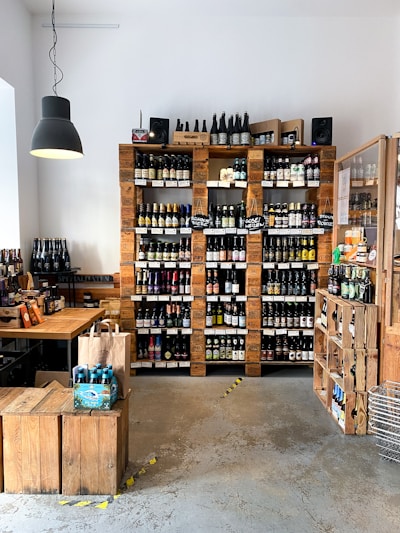 A quaint shop interior showcasing an extensive collection of bottled beverages, predominantly beer, arranged neatly on wooden shelves against a white wall. The rustic aesthetic is complemented by a large black hanging lamp, a wooden table, and crates that serve as both storage and decor. Several brown paper bags and packaging with vibrant labels are visible on the table.
