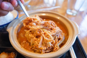 A clay pot filled with a spicy noodle and vegetable dish, being stirred with a spoon. Surrounding the pot are wooden chopsticks and a bowl of rice on a table with glasses in the background.