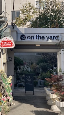 An outdoor archway with a sign on the top that says 'on the yard' with a tennis ball logo. Below and to the left is a Stella Artois sign. In the background, there is a blackboard sign that says 'SOLD OUT' with some other text underneath. There are plants and trees visible through the archway, and several flower pots and floral decorations line the sides of the path.