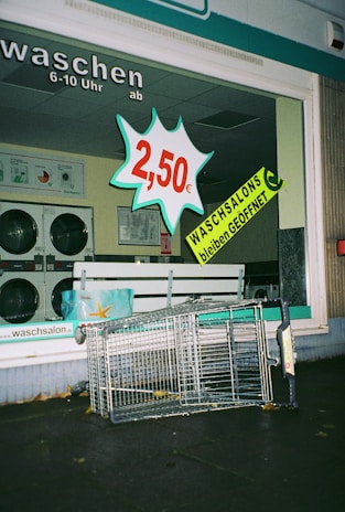 A laundromat display window features a bold price sign for 2.50 euros alongside a prominent announcement that laundromats remain open, both in bright and attention-grabbing colors. Inside, washing machines can be seen. Outside the laundromat, a toppled shopping cart lies on the pavement.