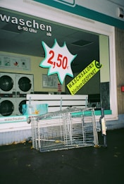 A laundromat display window features a bold price sign for 2.50 euros alongside a prominent announcement that laundromats remain open, both in bright and attention-grabbing colors. Inside, washing machines can be seen. Outside the laundromat, a toppled shopping cart lies on the pavement.