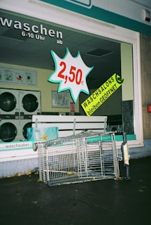 A laundromat display window features a bold price sign for 2.50 euros alongside a prominent announcement that laundromats remain open, both in bright and attention-grabbing colors. Inside, washing machines can be seen. Outside the laundromat, a toppled shopping cart lies on the pavement.