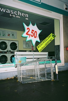A laundromat display window features a bold price sign for 2.50 euros alongside a prominent announcement that laundromats remain open, both in bright and attention-grabbing colors. Inside, washing machines can be seen. Outside the laundromat, a toppled shopping cart lies on the pavement.
