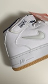 A white sneaker featuring a gum sole and detailed stitching. The shoe has the letters 'NYC' embroidered on the strap and a swoosh design in white on the side. A red tag with white text is attached to the shoe.