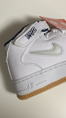 A white sneaker featuring a gum sole and detailed stitching. The shoe has the letters 'NYC' embroidered on the strap and a swoosh design in white on the side. A red tag with white text is attached to the shoe.