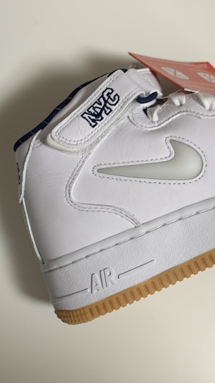 A white sneaker featuring a gum sole and detailed stitching. The shoe has the letters 'NYC' embroidered on the strap and a swoosh design in white on the side. A red tag with white text is attached to the shoe.