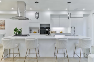 A modern, white kitchen featuring a large island with four quilted bar stools. Sleek cabinetry lines the walls, and stainless steel appliances are integrated seamlessly. Two pendant lights hang above the island, which is adorned with wine bottles and a bouquet of flowers. A built-in stovetop with an overhead vent is present. A potted plant adds a touch of greenery, and a piece of wall art is partially visible.