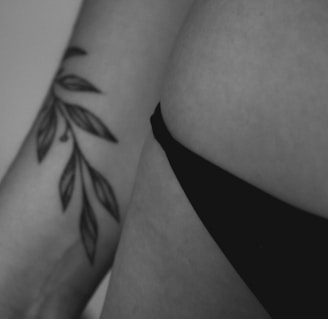 A close-up black and white photograph focusing on a person's hip and arm. The person is wearing a black undergarment, and their arm has a tattoo of leaves.