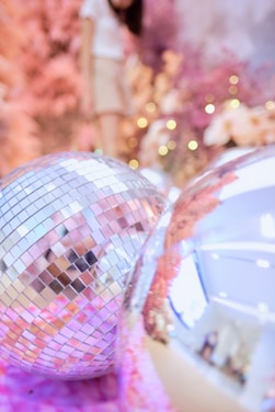 Large reflective disco balls are situated in the foreground, creating a festive atmosphere. The background features a woman standing with vibrant pink and purple floral decorations along with small twinkling lights, which enhance the celebratory feel.