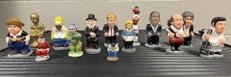 A collection of small, colorful figurines depicting various pop culture and historical characters are arranged in two rows on a flat surface. Each figurine is uniquely styled to represent their iconic features.