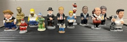 A collection of small, colorful figurines depicting various pop culture and historical characters are arranged in two rows on a flat surface. Each figurine is uniquely styled to represent their iconic features.