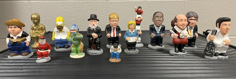 A collection of small, colorful figurines depicting various pop culture and historical characters are arranged in two rows on a flat surface. Each figurine is uniquely styled to represent their iconic features.