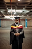 A person wearing a graduation cap and gown stands in a parking garage. The gown is black with a maroon and yellow hood or stole. The person has a serious expression and is dressed in formal attire with a tie and watch.