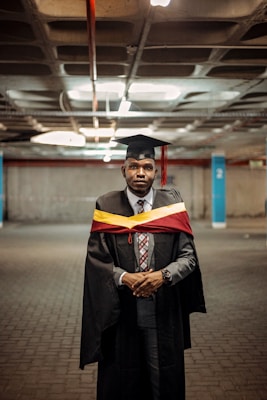 A person wearing a graduation cap and gown stands in a parking garage. The gown is black with a maroon and yellow hood or stole. The person has a serious expression and is dressed in formal attire with a tie and watch.
