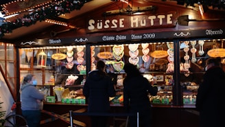 A festive market stall decorated with Christmas lights and evergreen garlands. The stall displays a variety of sweets, prominently featuring heart-shaped gingerbread cookies with icing. Customers are gathered at the counter making purchases, with some wearing face masks.