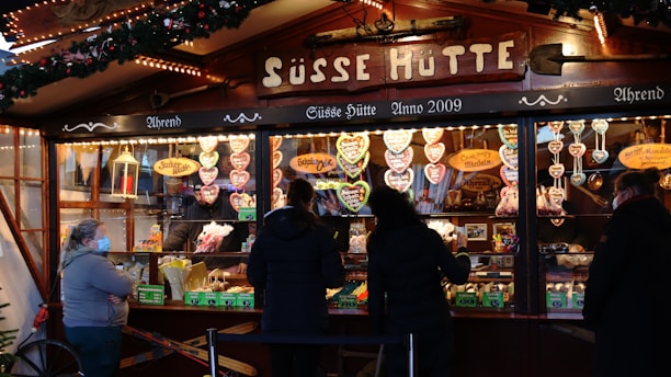 A festive market stall decorated with Christmas lights and evergreen garlands. The stall displays a variety of sweets, prominently featuring heart-shaped gingerbread cookies with icing. Customers are gathered at the counter making purchases, with some wearing face masks.