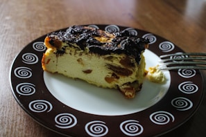 A slice of dessert with a browned top and visible raisins is placed on a decorative plate with spiral patterns. The plate is on a wooden surface, and a fork with a bit of dessert on its prongs rests beside the slice.