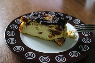 A slice of dessert with a browned top and visible raisins is placed on a decorative plate with spiral patterns. The plate is on a wooden surface, and a fork with a bit of dessert on its prongs rests beside the slice.