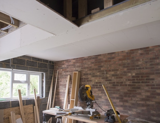 A partially renovated room with a brick wall and an unfinished ceiling. A window allows natural light to enter, and various construction materials are scattered around. A circular saw is placed on a workbench surrounded by planks and boards.