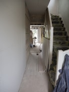 A hallway under construction with exposed brick walls, insulation on the stairs, and scattered construction materials. There are boards against the walls and tools are visible in the distance. The space is mostly unfinished and appears to be in the mid-stages of renovation.