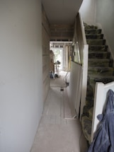 A hallway under construction with exposed brick walls, insulation on the stairs, and scattered construction materials. There are boards against the walls and tools are visible in the distance. The space is mostly unfinished and appears to be in the mid-stages of renovation.