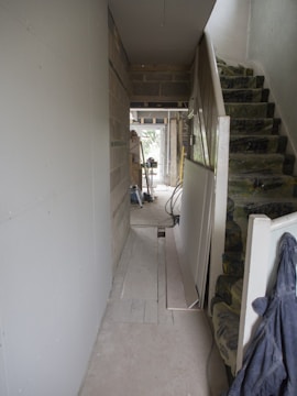 A hallway under construction with exposed brick walls, insulation on the stairs, and scattered construction materials. There are boards against the walls and tools are visible in the distance. The space is mostly unfinished and appears to be in the mid-stages of renovation.