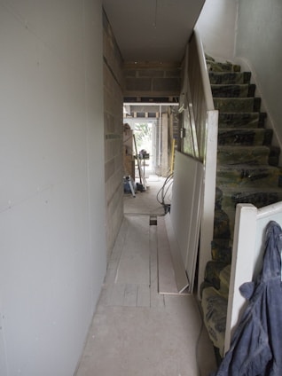 A hallway under construction with exposed brick walls, insulation on the stairs, and scattered construction materials. There are boards against the walls and tools are visible in the distance. The space is mostly unfinished and appears to be in the mid-stages of renovation.