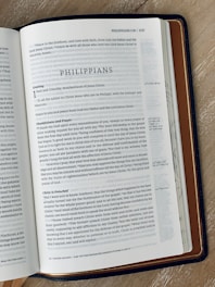 An open book showing a page from the Bible. The page is titled 'Philippians' and includes text with headings such as 'Greeting' and 'Thankfulness and Prayer'. The book is lying on a wooden surface, and it has a dark brown leather cover.