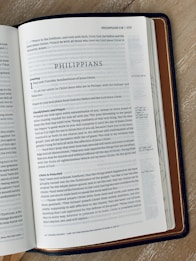 An open book showing a page from the Bible. The page is titled 'Philippians' and includes text with headings such as 'Greeting' and 'Thankfulness and Prayer'. The book is lying on a wooden surface, and it has a dark brown leather cover.