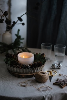 A candle sits in the center of a festive arrangement surrounded by pinecones and greenery. Two empty glasses are nearby, along with a spool of twine, scissors, and metallic bells. A wooden star and some branches add to the rustic decor. The overall setup rests on a table covered with a light cloth.