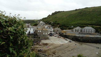 A quaint coastal village with stone and brick houses tightly packed together. The village is surrounded by lush green hills, and there is a sandy area in the forefront, possibly a beach or harbor. A few people are visible walking or sitting, and a large plant is seen in the left corner.