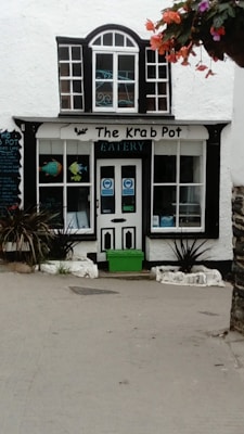 A quaint eatery named 'The Krab Pot' is seen with a charming facade featuring a black and white color scheme. Above the door and windows, the name is displayed with decorative elements like a small crab image and aquatic-themed graphics. The windows have a decorative grid, and there is a green planter box near the entrance. Bright flowers are hanging from above, adding a splash of color.