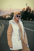 A person is standing outdoors wearing a beige hoodie under a white vest with 'Balenciaga' written on it. They are also wearing a beige cap and large black sunglasses. Background includes a road and trees under a muted, overcast sky.