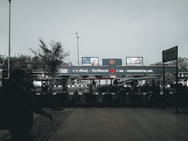 A public transport area with a metro station sign in both Hindi and English for SBI - VISHWAVIDYALAYA. Several auto rickshaws are parked in front of the station. There are billboards above the station, and a few trees and lamp posts are visible. A silhouette of a person walking covers part of the foreground.