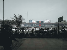 A public transport area with a metro station sign in both Hindi and English for SBI - VISHWAVIDYALAYA. Several auto rickshaws are parked in front of the station. There are billboards above the station, and a few trees and lamp posts are visible. A silhouette of a person walking covers part of the foreground.