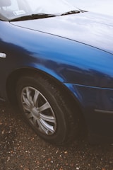 A close-up view of a car's front wheel and part of the body. The car is painted in a glossy blue finish, and the wheel features a metallic alloy rim. The surface of the car reflects light, showing some reflections of the sky and surroundings. The vehicle is parked on a gravel surface with various colors of small stones and pebbles visible.