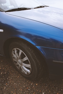 A close-up view of a car's front wheel and part of the body. The car is painted in a glossy blue finish, and the wheel features a metallic alloy rim. The surface of the car reflects light, showing some reflections of the sky and surroundings. The vehicle is parked on a gravel surface with various colors of small stones and pebbles visible.