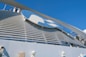 A large structure featuring a prominent logo with a blue and white color scheme is set against a clear blue sky. The design includes a stylized profile of a person with flowing hair. The image also includes industrial elements such as pipes and vents.
