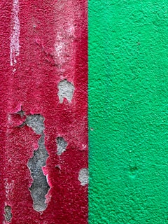 The image features a close-up view of a textured wall that is divided into two halves. The left side has a red paint that is peeling off, revealing patches of gray beneath. The right side displays a solid green paint with a rough texture.