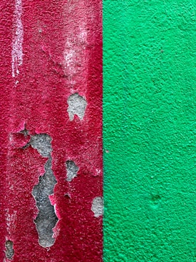 The image features a close-up view of a textured wall that is divided into two halves. The left side has a red paint that is peeling off, revealing patches of gray beneath. The right side displays a solid green paint with a rough texture.