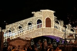 A bridge is beautifully decorated with numerous small white lights, creating a festive and enchanting atmosphere. Below the bridge, a crowd of people is gathered, surrounded by illuminated trees and structures.