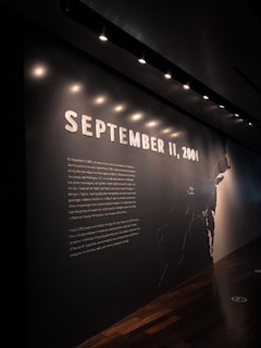 A dimly lit wall with the large embossed text 'September 11, 2001' prominently displayed, accompanied by a detailed narrative about the September 11 attacks. Above, small spotlights cast a soft light downwards, creating a solemn ambiance. The wall also features a map outline on the right side, partially illuminated.